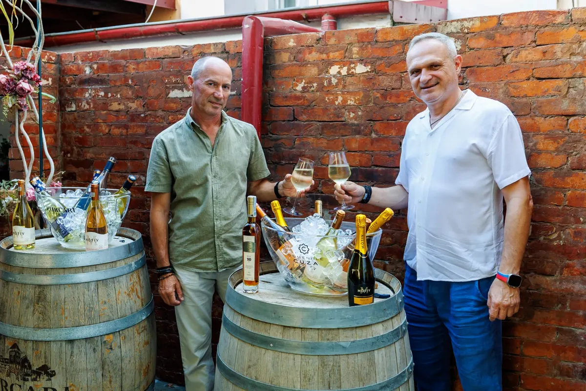 Doi bărbați cu pahare de vin alb, în fața butoaielor cu sticle și frapiere pline de gheață
