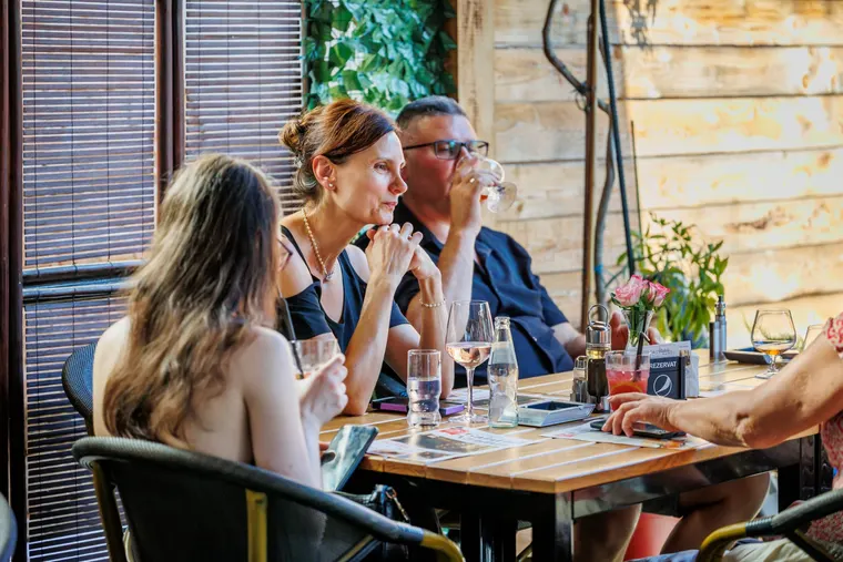 Trei oaspeți la masă, cu pahare de vin în față, lângă fereastra cu jaluzele de lemn
