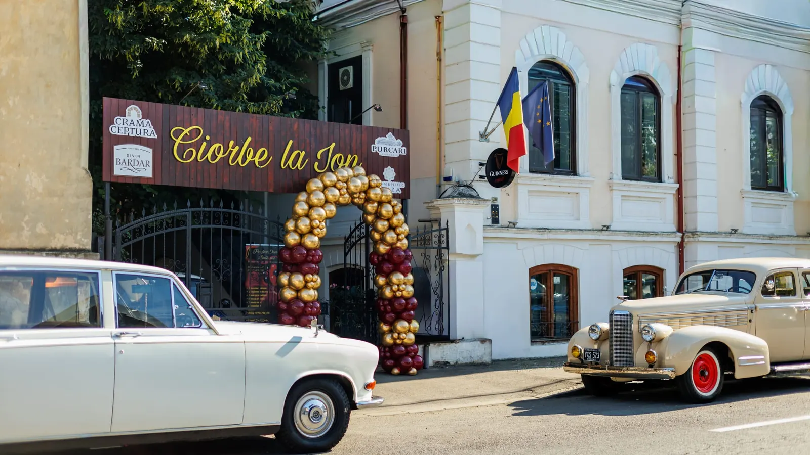 Intrarea restaurantului Ciorbe La Ion, cu firma de lemn și o arcadă de baloane aurii și vișinii la poartă, între două automobile de epocă oprite la bordură
