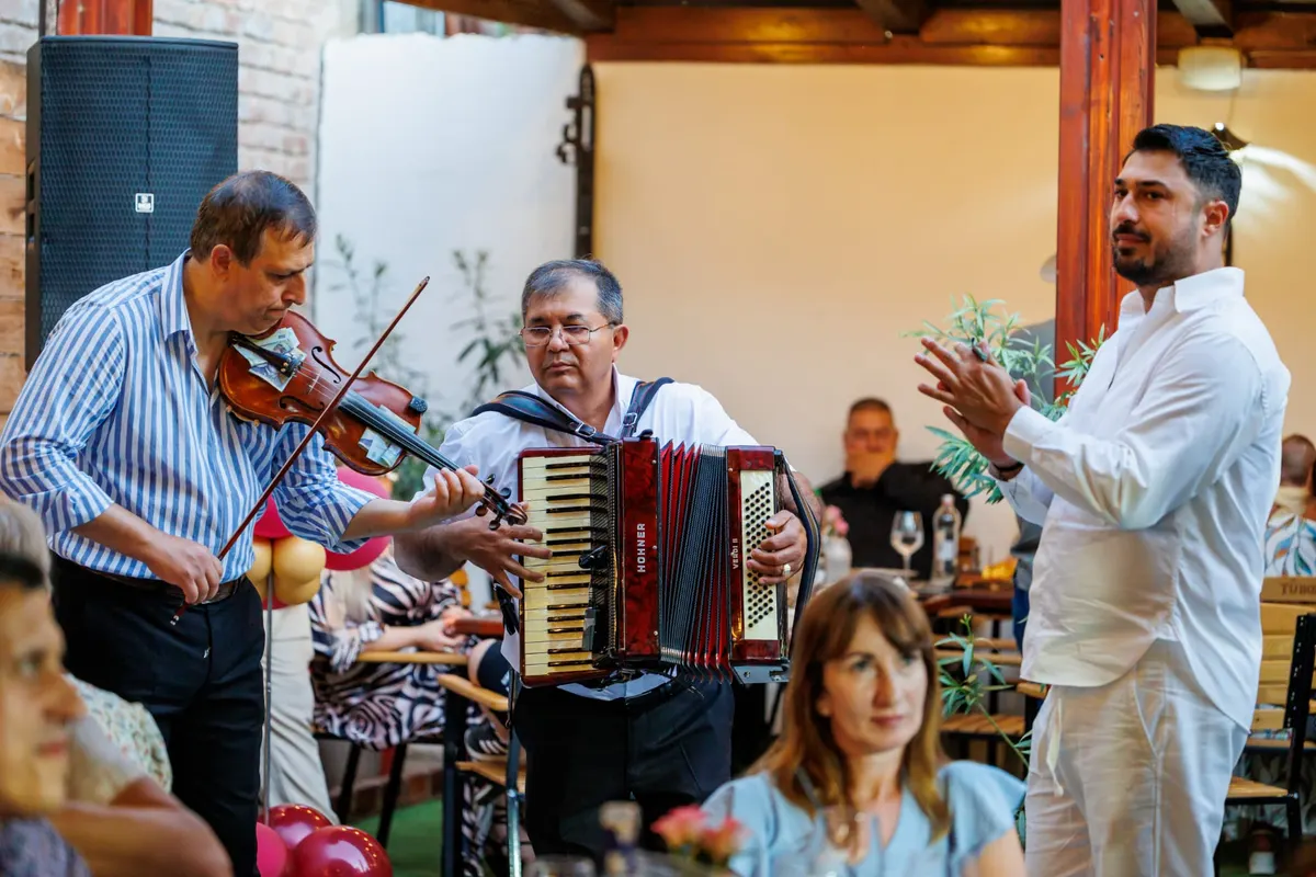 Vioară și acordeon cântate între mese, cu un oaspete care bate din palme alături