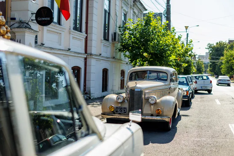 Două automobile de epocă oprite pe stradă, lângă o clădire veche, cu mașini obișnuite în spatele lor