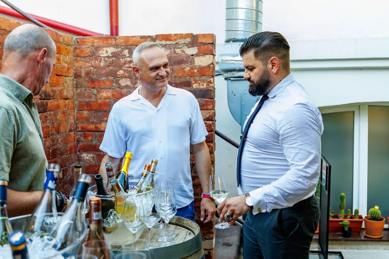 Trei bărbați în jurul unui butoi acoperit cu pahare și sticle, lângă un perete de cărămidă