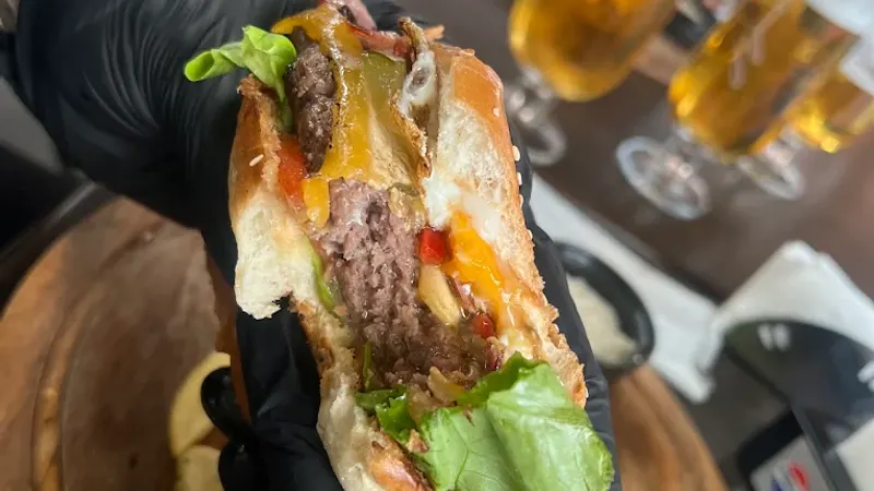 Burger tăiat în două, ținut deasupra unui platou de lemn, cu halbe de bere în spate