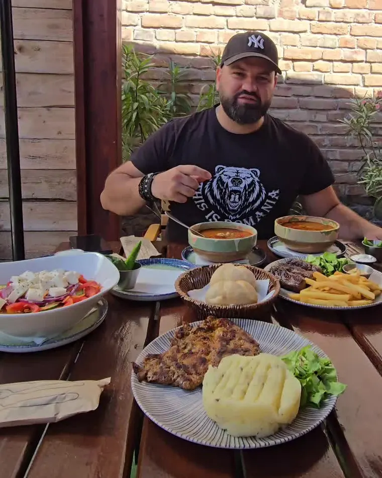 Ion la masă pe terasă, cu două străchini de ciorbă, friptură, cartofi prăjiți și mămăliguță
