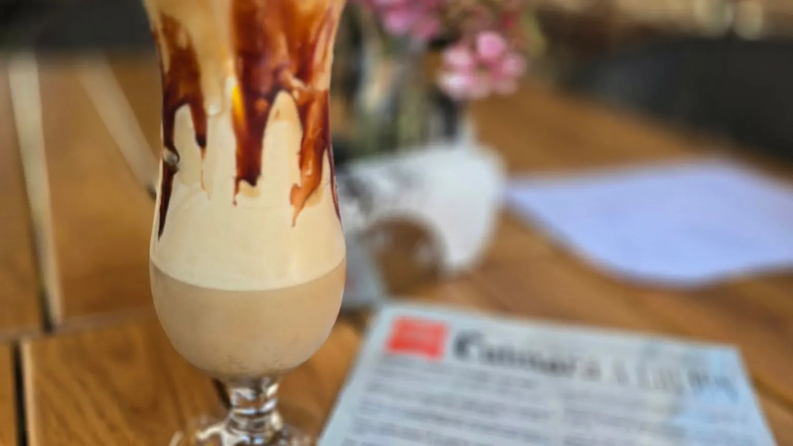 Pahar înalt de cafea cu frișcă și sirop de ciocolată, pe o masă de lemn